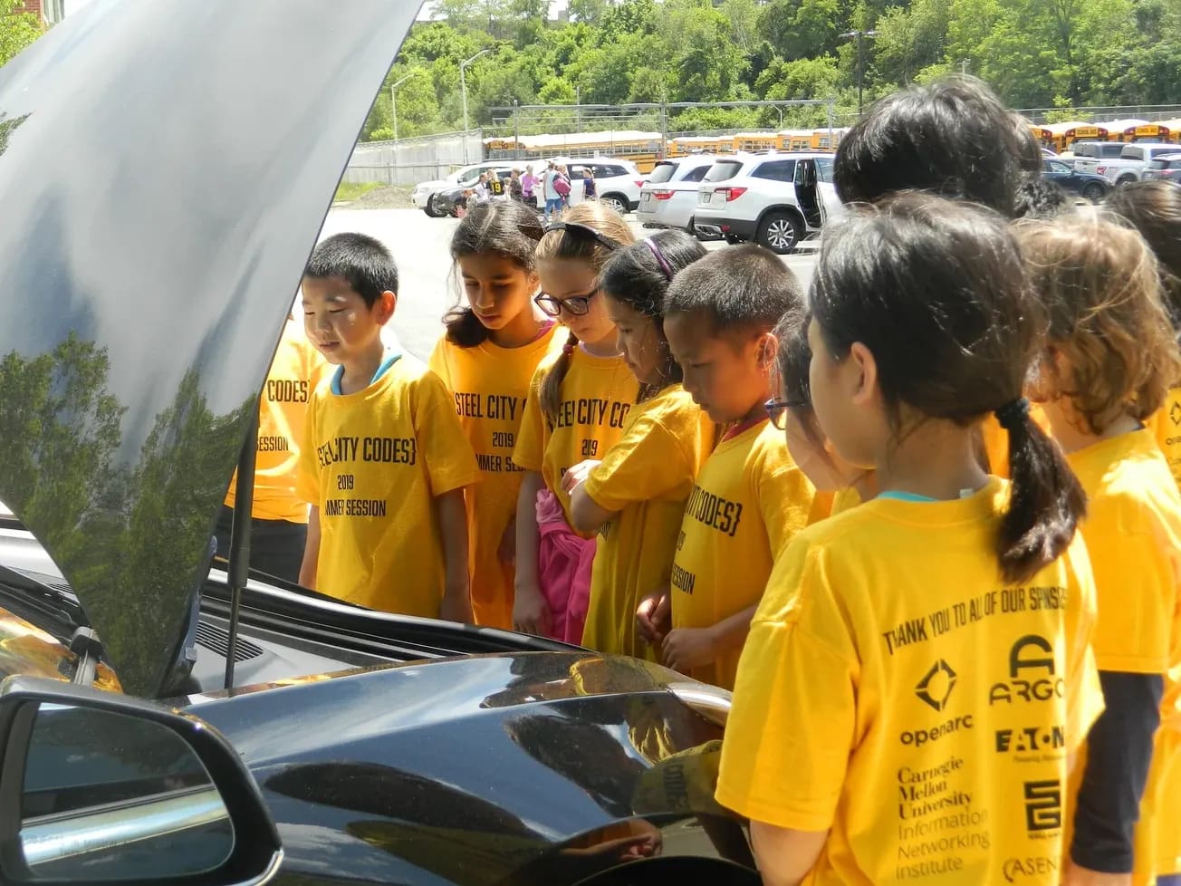 Campers in yellow Steel City Codes shirts printed with sponsor logos, looking under the open hood of a car during summer camp.