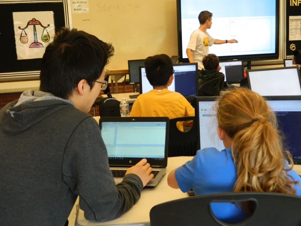 A mentor pointing at code on a large screen while students follow along on laptops.