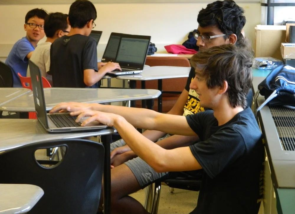 A high school mentor leaning in to help a younger student with code on a laptop.