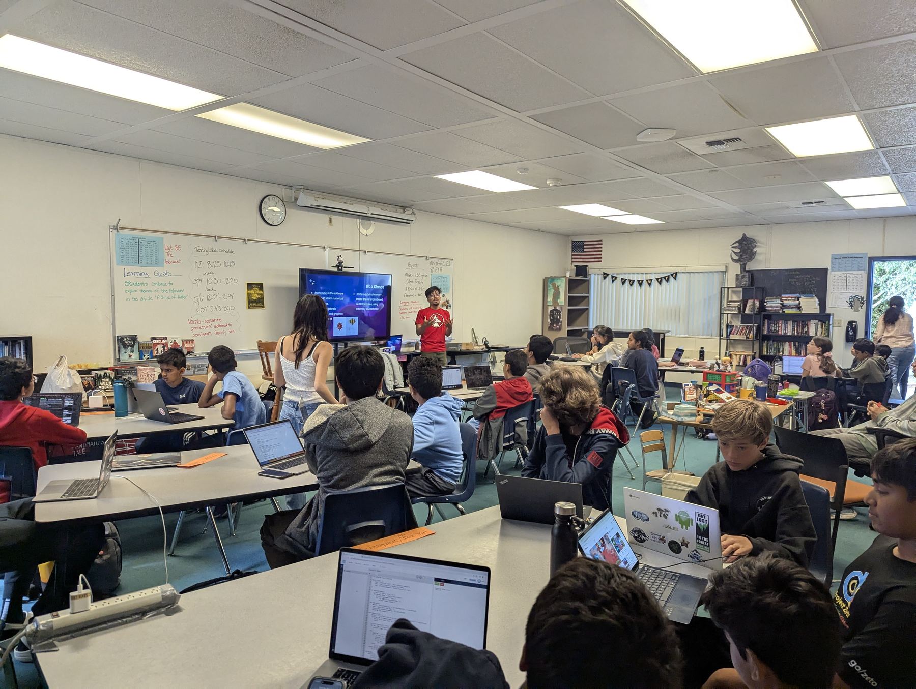 A high school mentor presenting a lesson at the front of a classroom while students follow along on laptops.