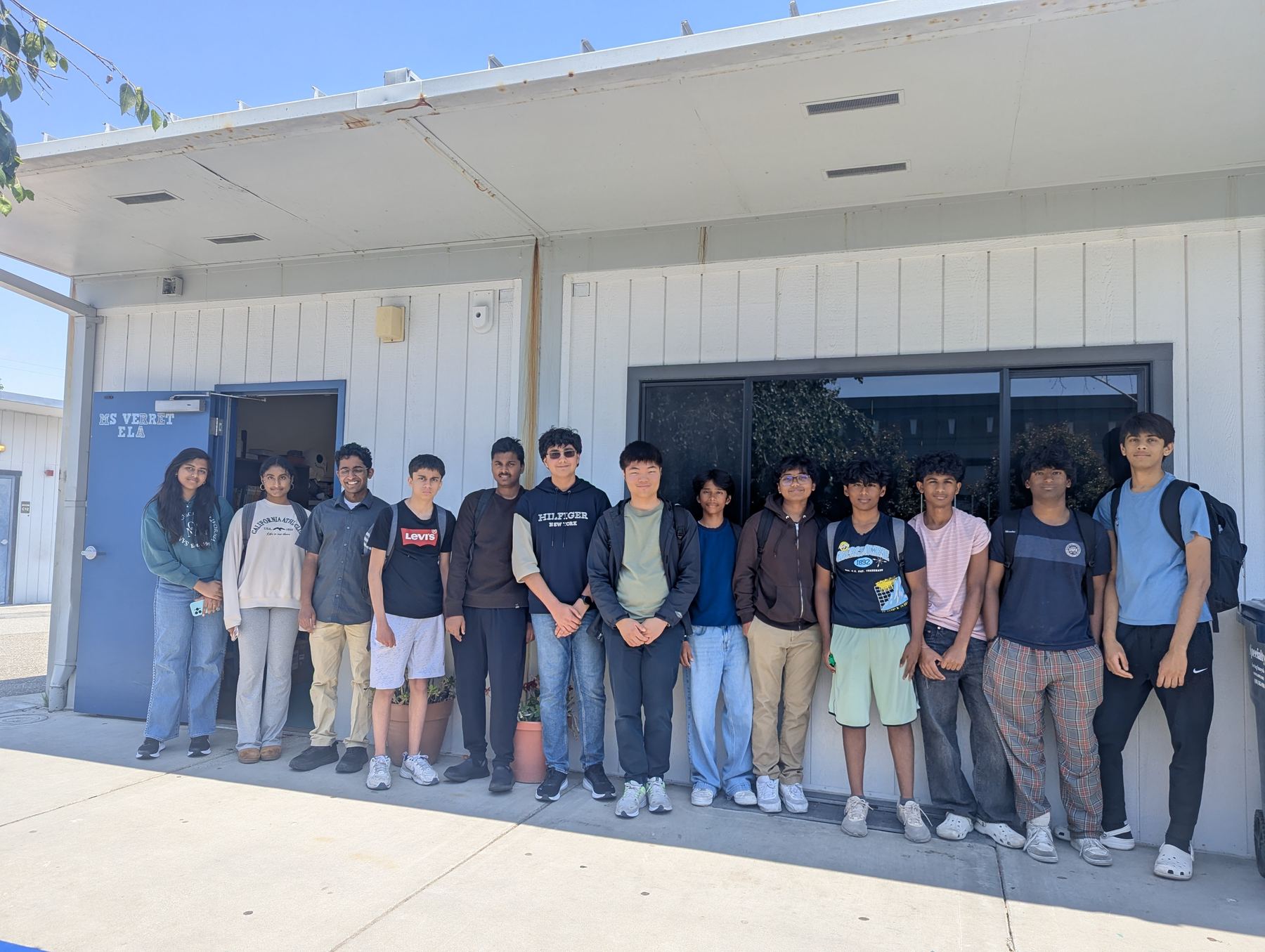 A line of high school volunteers standing together outside a classroom building on a sunny day.