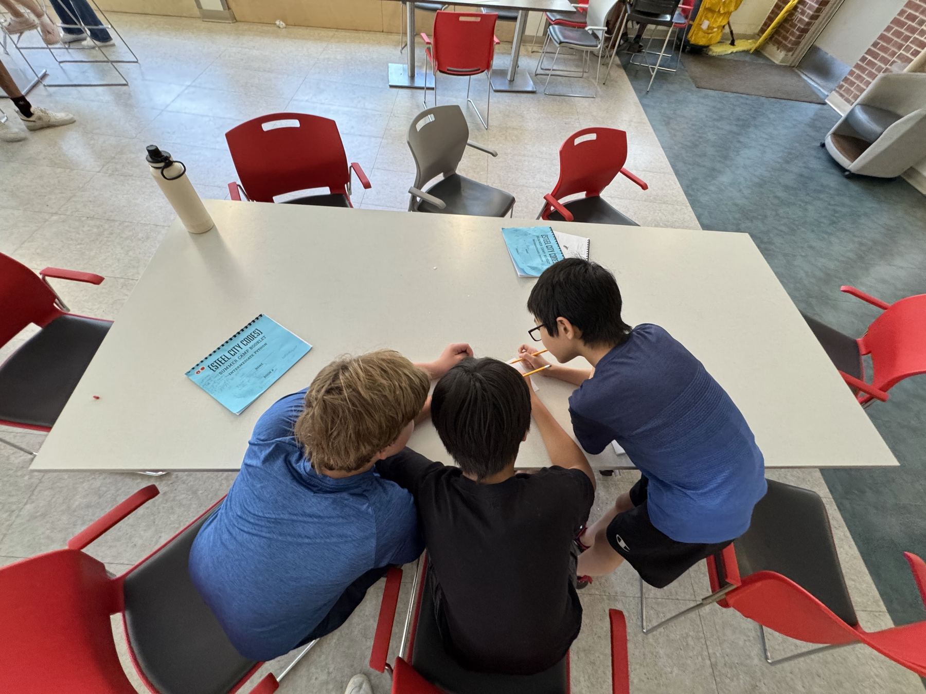 Three students working through a Steel City Codes worksheet together at a table, seen from above.