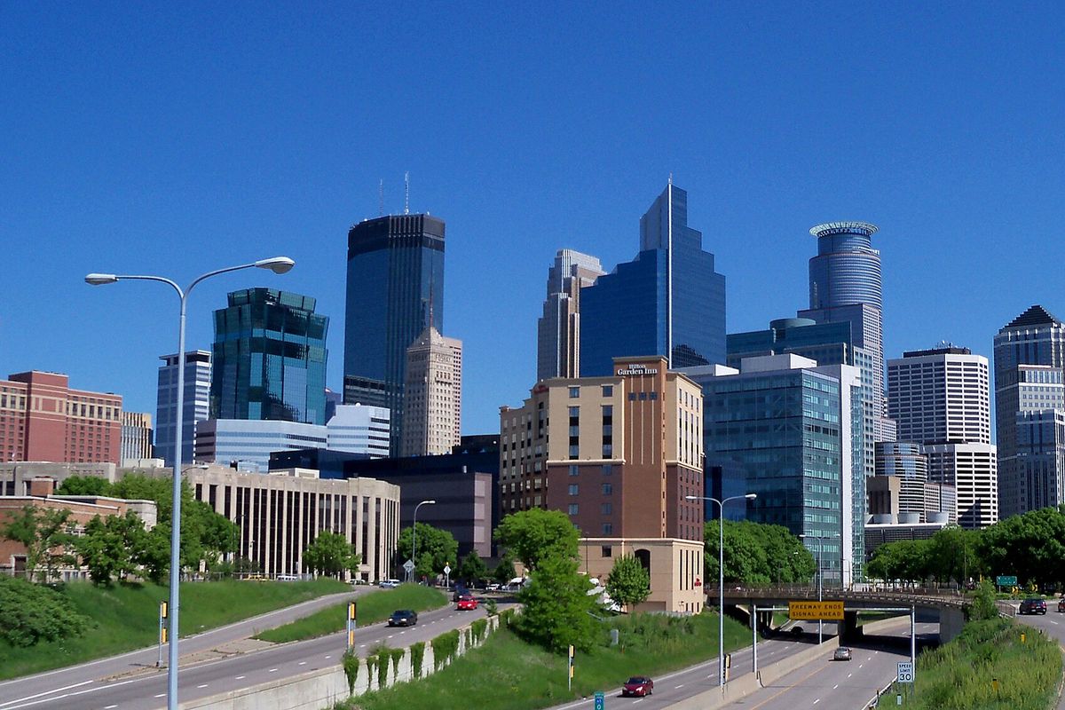 Downtown Minneapolis towers above green embankments and roads under a clear blue sky.